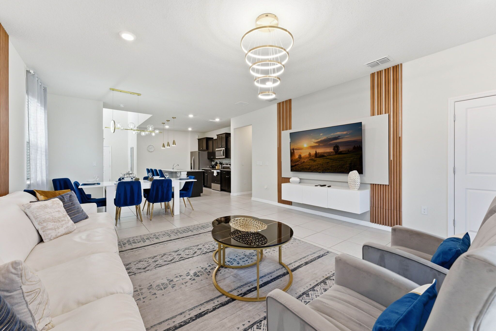 A modern living room with a sofa, coffee table, and TV. There is a dining area with chairs and a table. The room has a kitchen in the background.