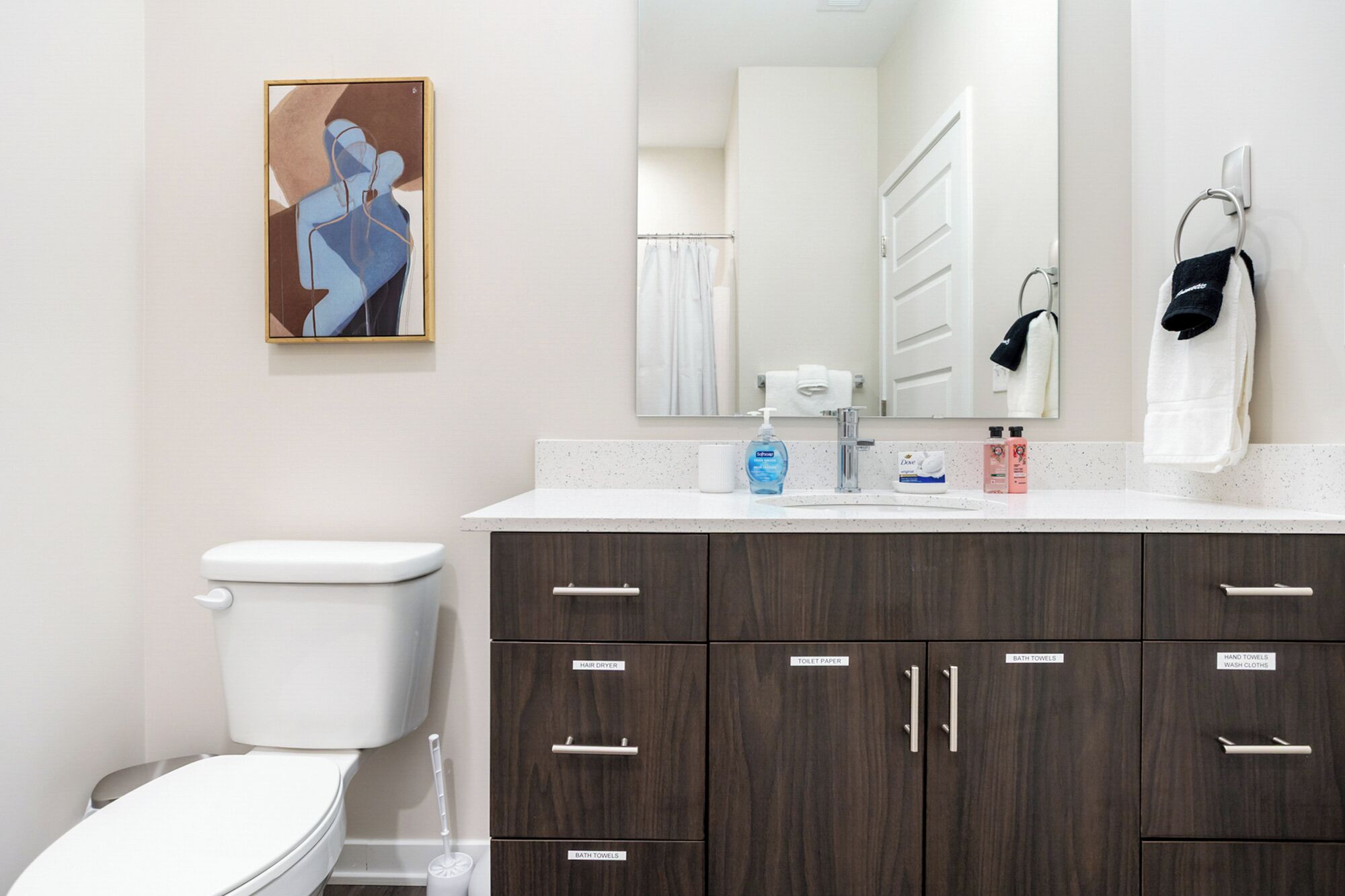 Ein modernes Badezimmer mit Toilette, Waschbecken mit Spiegel und einem Schrank mit beschrifteten Schubladen für Haartrockner, Toilettenpapier, Badetücher und Handtücher.