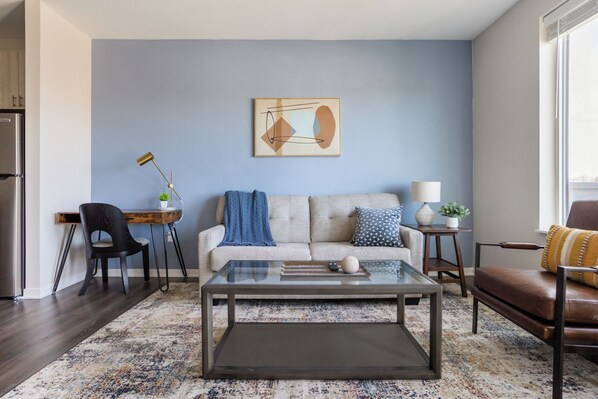 A living room with a sofa, coffee table, and chairs, a desk with a lamp, and a painting on the wall.