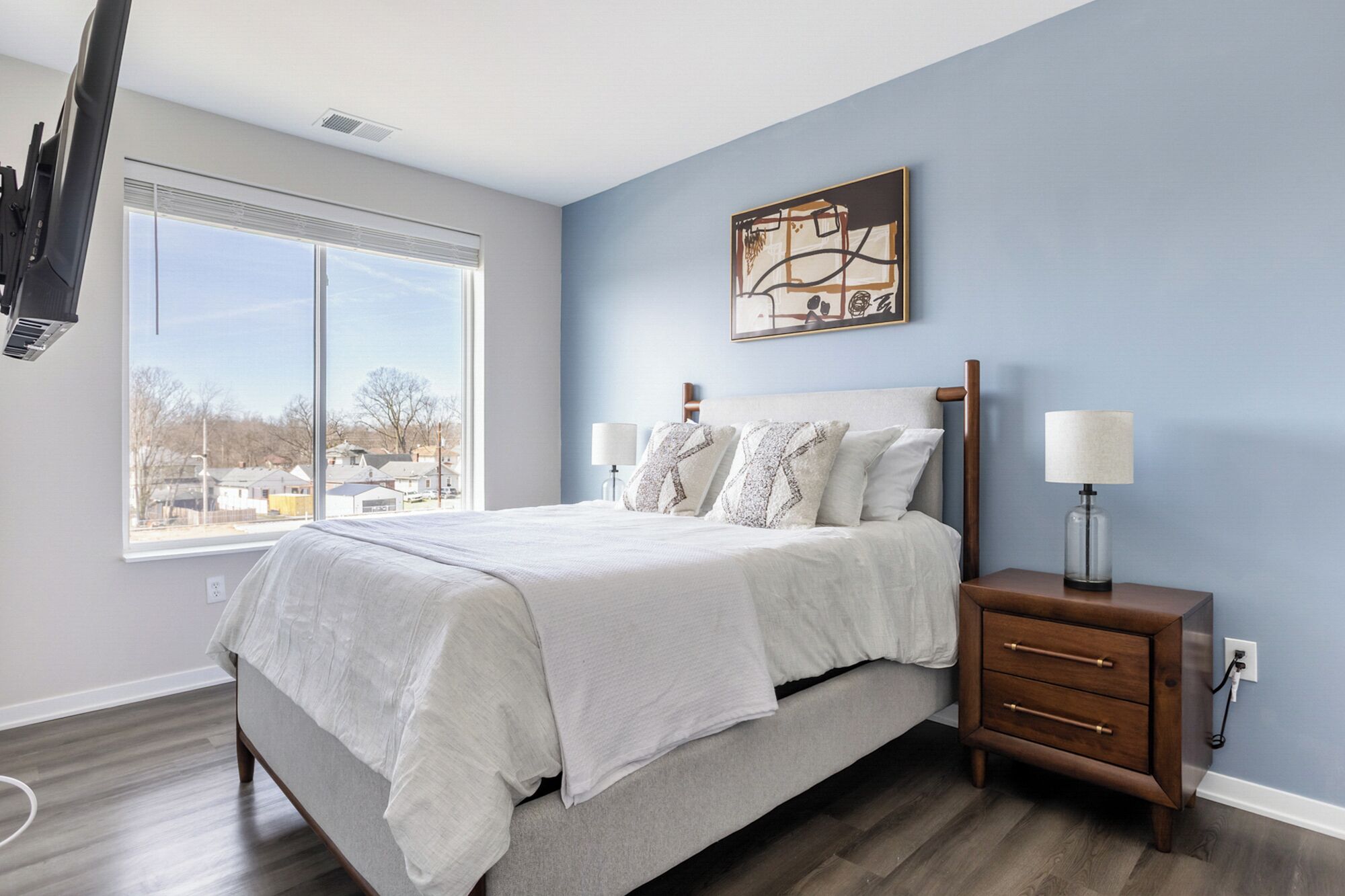 A bedroom with a large bed, bedside lamps, a nightstand, a window with a view, and a painting on the wall.