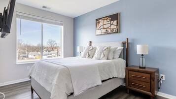 A bedroom with a large bed, bedside lamps, a nightstand, a window with a view, and a painting on the wall.