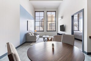A modern living room with a round wooden table, a sofa, a TV, and large windows offering a city view. - Landing Apartments near Connecticut State Capitol (Hartford)