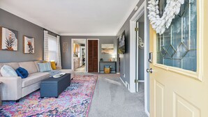 Een woonkamer met een bank, voetenbankje en een kleurrijk vloerkleed. Er is een deur versierd met een krans en een raam met een geometrisch patroon.