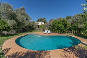 Una piscina con borde de baldosas, rodeada de árboles y un muro de piedra.