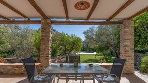 Eine überdachte Terrasse mit Tisch und Stühlen, Steinpfeilern und Blick auf ein Schwimmbad und Grünflächen.