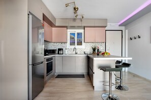 A modern kitchen with stainless steel appliances, a central island with bar stools, and a window with a view. - South Blue Beach & Park Apartment in Vouliagmeni (Vouliagmeni)
