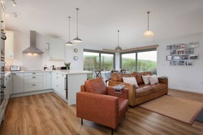 A modern kitchen with white cabinets, a dining area with a brown leather sofa, and a view of the outdoors through large windows. - Sloopside - The Annexe, Bantham, South Devon (Bantham)