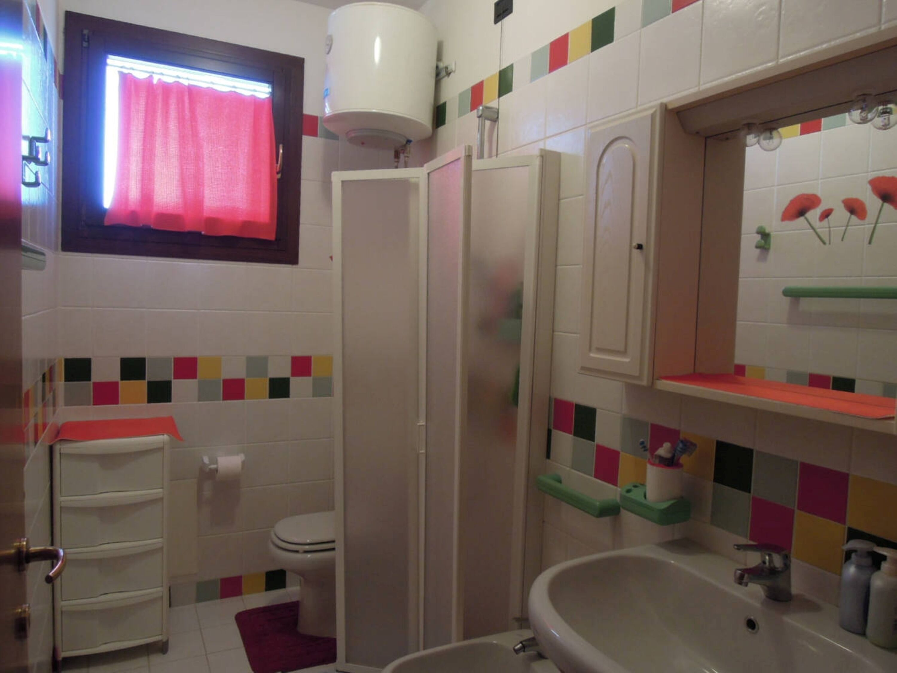 A bathroom with a toilet, a shower, a sink, and a window with a red curtain.
