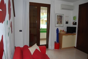 A living room with a red sofa, a television on a wooden cabinet, and a sliding glass door leading to a balcony. - Relaxing Flat With Pool in a Quiet Holiday Village (San Michele al Tagliamento)