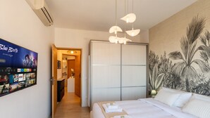 A hotel room with a bed, a wall-mounted TV displaying a movie, a wardrobe, and a tropical-themed wall mural. - GuestHost - Modern Flat with Lovely Terrace (Sanremo)