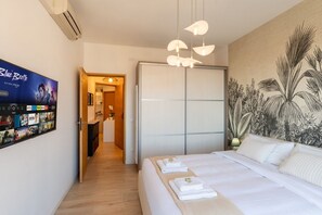 A hotel room with a bed, a wall-mounted TV displaying a movie, a wardrobe, and a tropical-themed wall mural. - GuestHost - Modern Flat with Lovely Terrace (Sanremo)