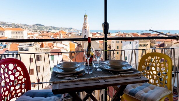 A balcony with a table set for two, a bottle, and glasses, overlooking a cityscape with buildings and a tower. - GuestHost - Modern Flat with Lovely Terrace (Sanremo)