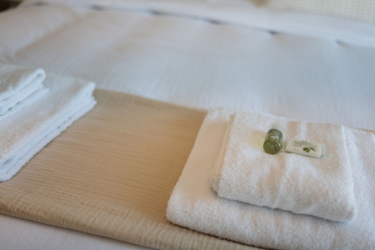 En prydligt vikt vit handduk på en beige matta, en hög med vita handdukar och en liten flaska med en etikett på en vit yta.