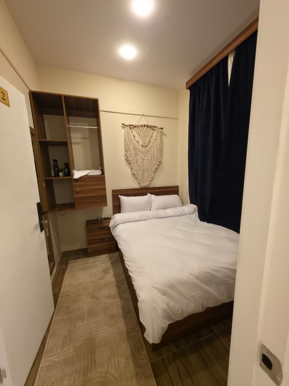 Een slaapkamer met een bed, een houten garderobe, een macramé wanddecoratie en een donker gordijn.