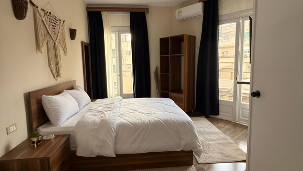 Une chambre à coucher avec un lit en bois, une table de chevet, une armoire et une grande fenêtre avec des rideaux.