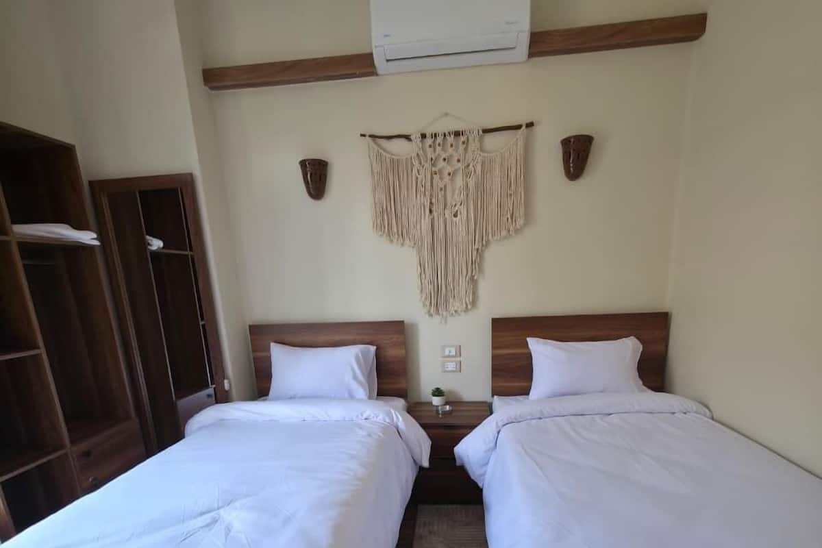 A hotel room with two beds, wooden headboards, a macrame wall hanging, an air conditioning unit, and a wooden wardrobe.