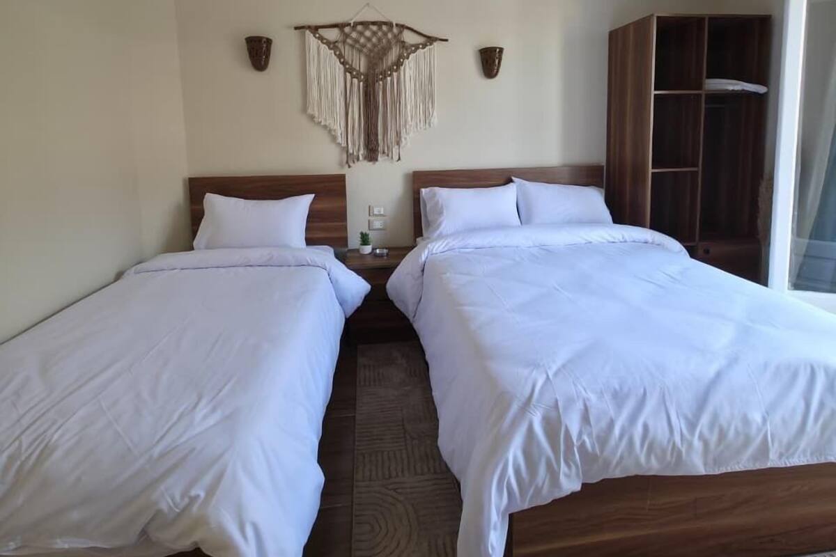 A hotel room with two beds, a wooden headboard, a wall-mounted air conditioner, a decorative macrame piece, and a wardrobe.