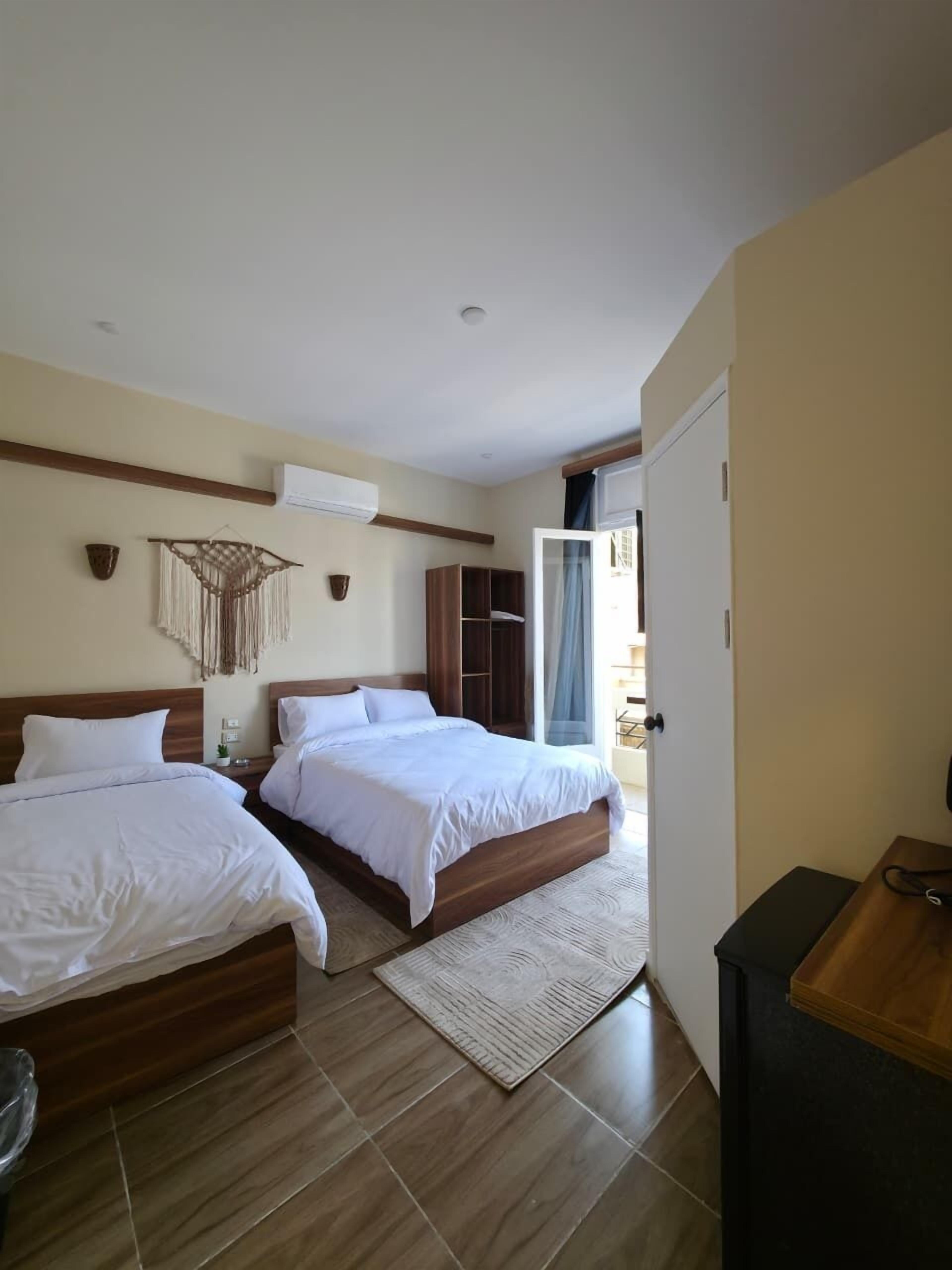 Hotellihuone, jossa on kaksi sänkyä, puinen vaatekaappi, pieni työpöytä ja näkymä ulos.