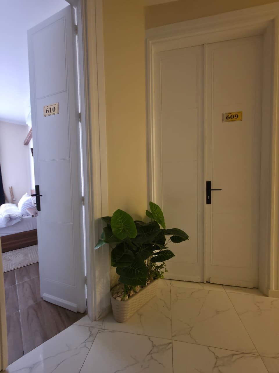 Een gang met twee deuren, waarvan één het nummer 610 heeft en de andere 609, die leidt naar een kamer met een bed en een potplant.