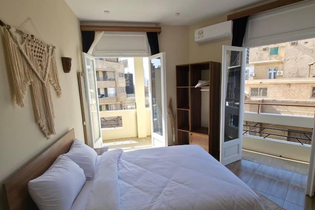 A bedroom with a bed, a wooden nightstand, a macrame wall hanging, and a view of the outside balcony.