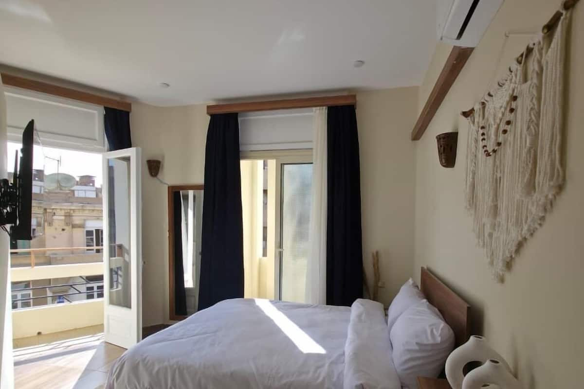 A bedroom with a large bed, a wooden headboard, a macrame wall hanging, and a view of the outside.
