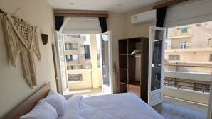 Une chambre avec un grand lit, une table de chevet en bois, une décoration murale en macramé et une vue sur le balcon extérieur.