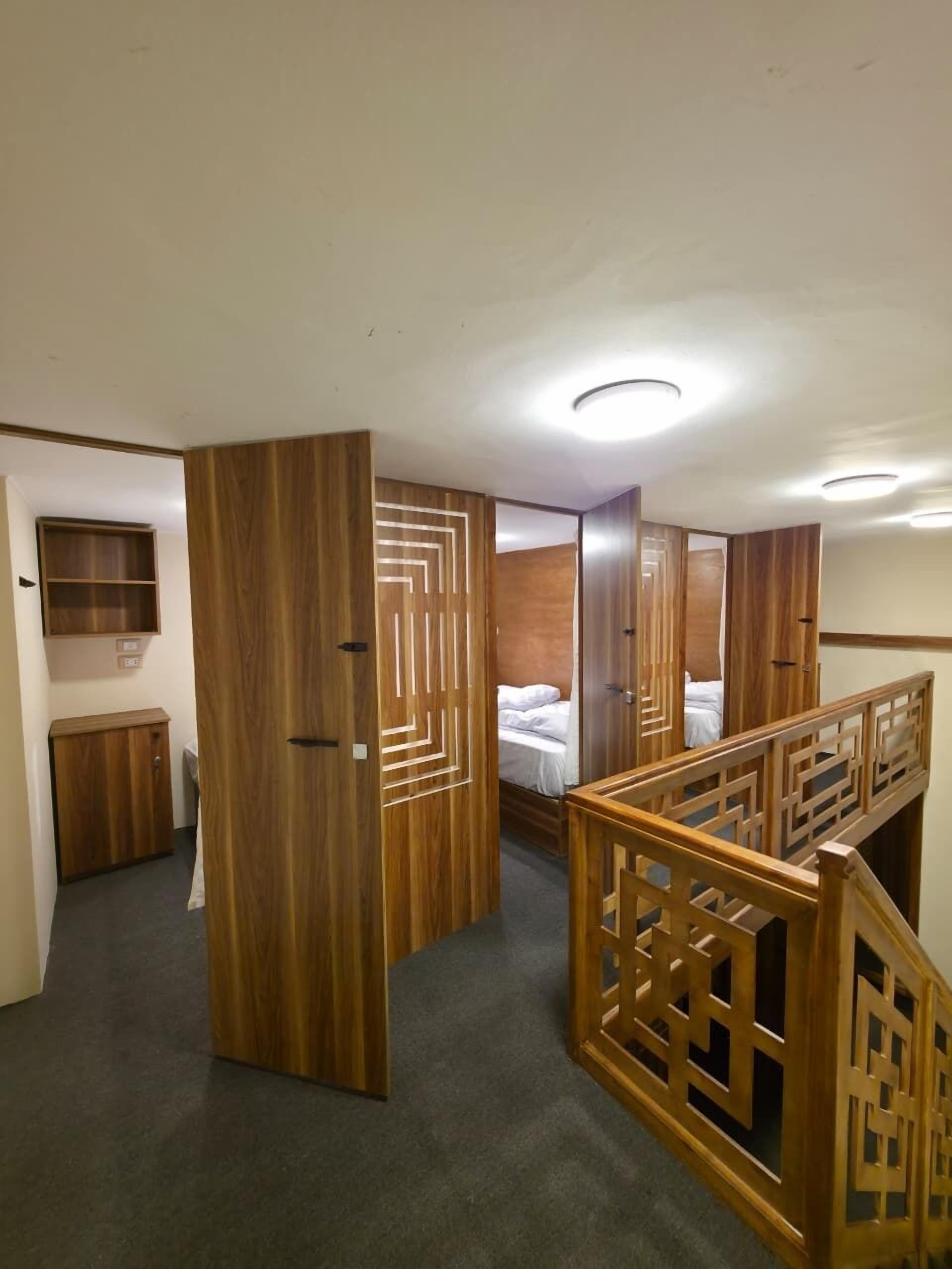 A hotel room with a wooden wardrobe, a bed, a small cabinet, and a staircase with a wooden railing.