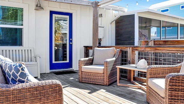 A covered outdoor deck with wicker furniture, a blue door, and a view of a house in the background. - 40th Street Cottage (Port St. Joe)