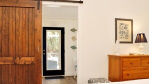 A room with a wooden sliding door, a white wall, a framed fish illustration, a lamp, and a wooden dresser. - 40th Street Cottage (Port St. Joe)
