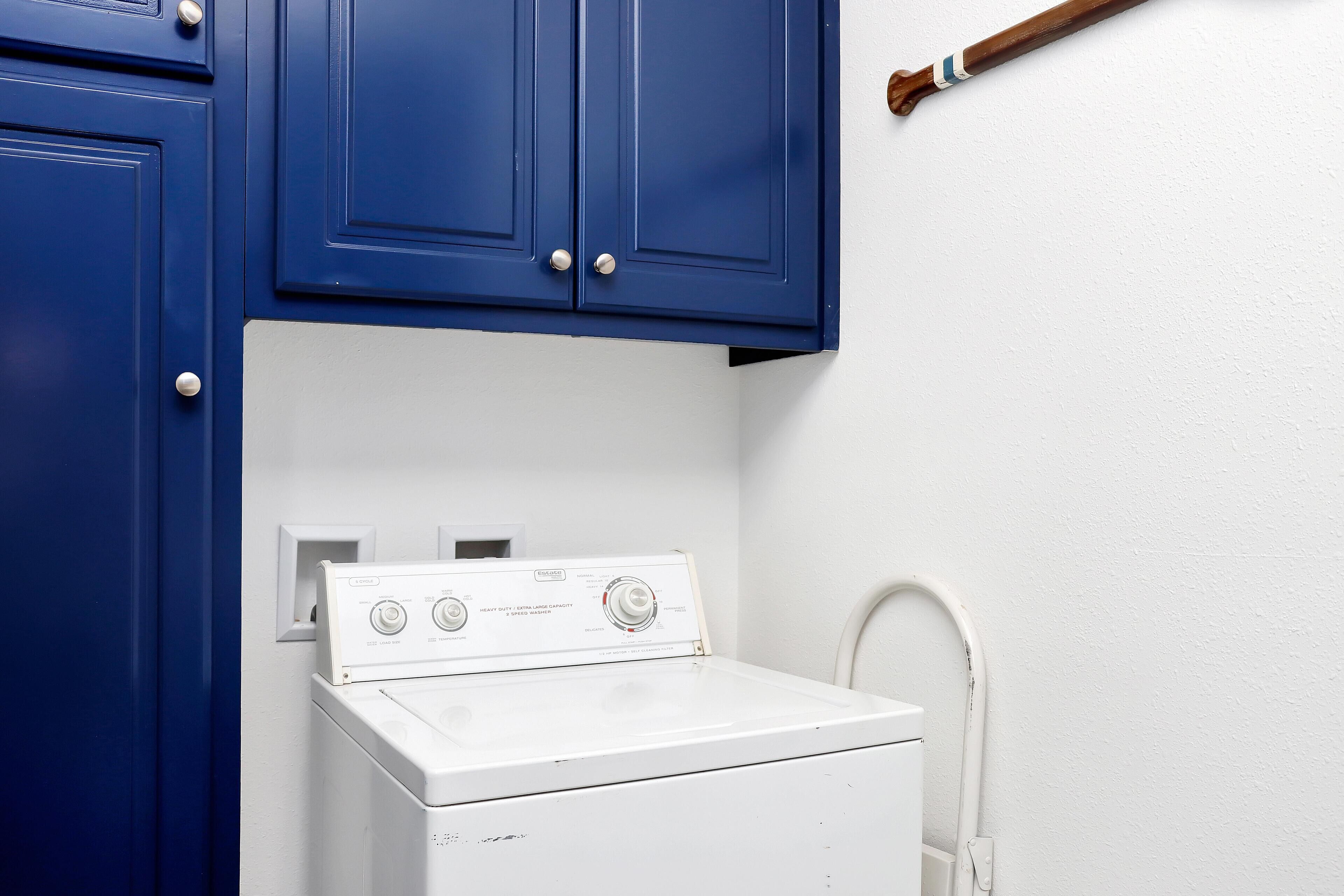 Une machine à laver blanche, placée sous des placards bleus, avec un mur blanc et une poignée en bois.