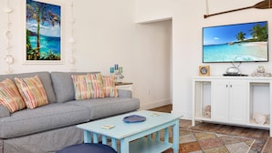 A living room with a grey sofa, colorful pillows, a blue coffee table, a TV on a white cabinet, and a painting of a beach scene. - 40th Street Cottage (Port St. Joe)