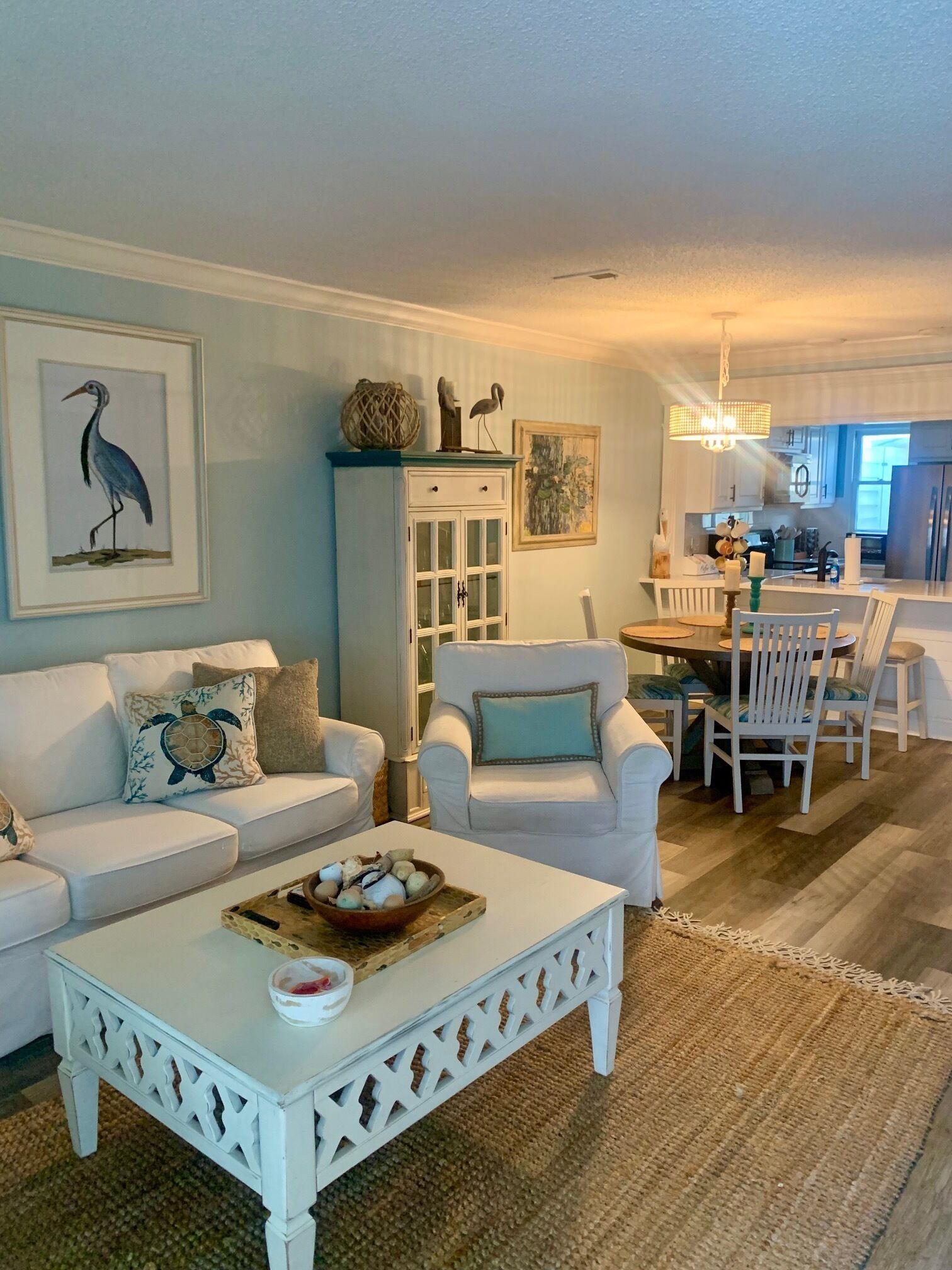 A living room with a white sofa, a white armchair, a coffee table with decorative items, a dining area with a table and chairs, and a kitchen in the background.