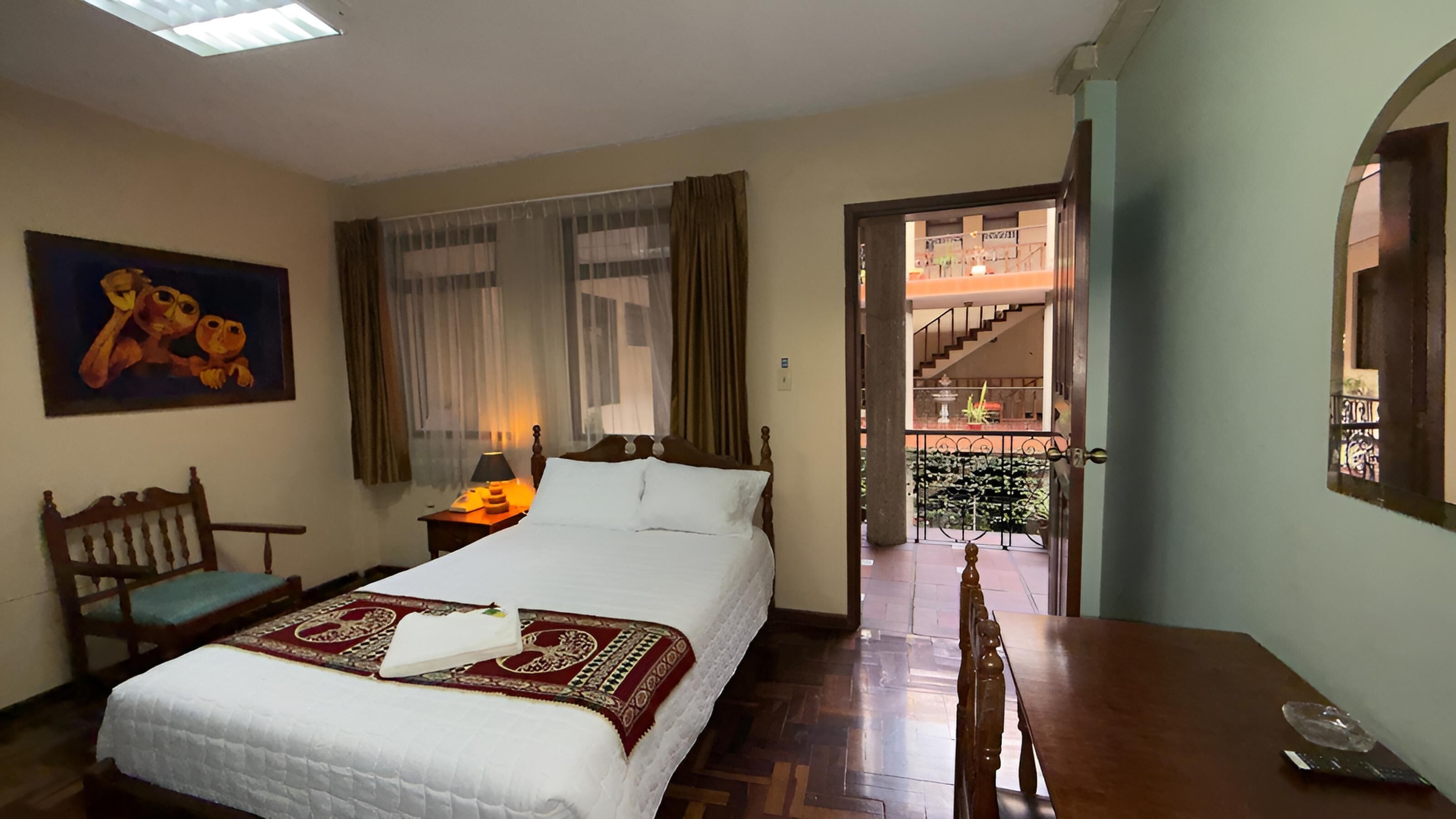 Une chambre d’hôtel comprenant un lit, un bureau, une chaise, un tableau, une fenêtre avec des rideaux et une vue sur le balcon extérieur.