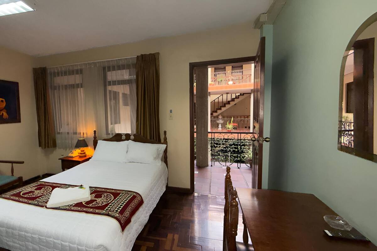 A hotel room with a bed, a desk, a chair, a painting, a window with curtains, and a view of the outside balcony.