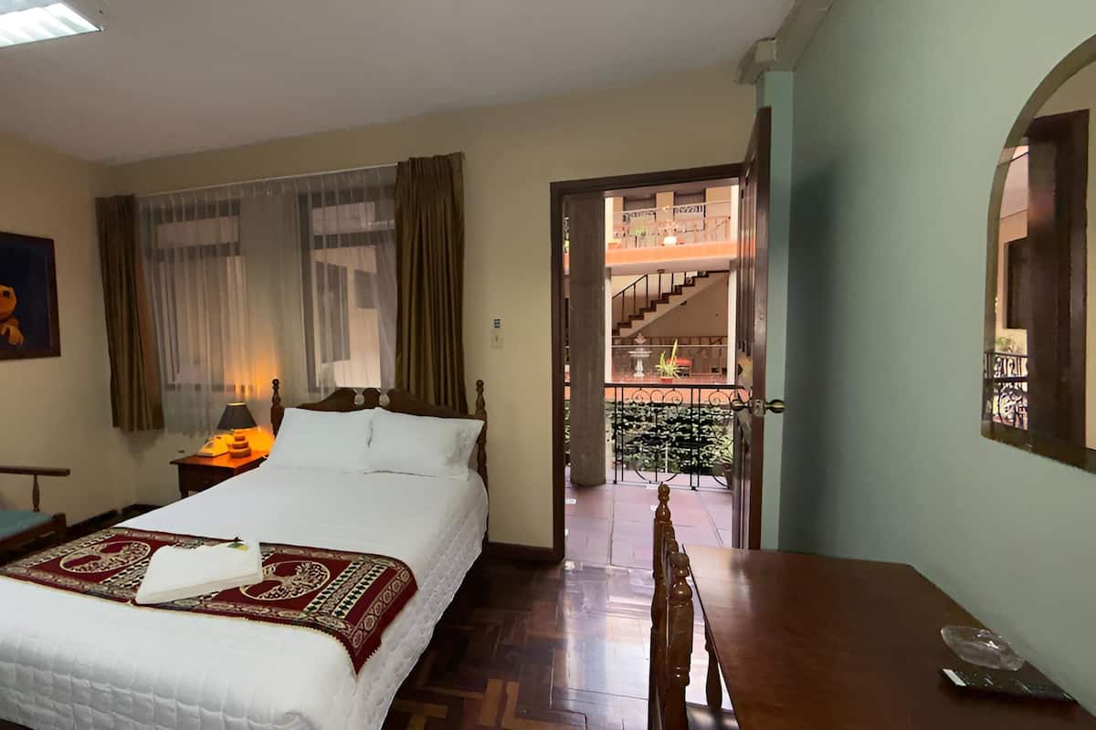 Une chambre d’hôtel comprenant un lit, un bureau, une chaise, un tableau, une fenêtre avec des rideaux et une vue sur le balcon extérieur.