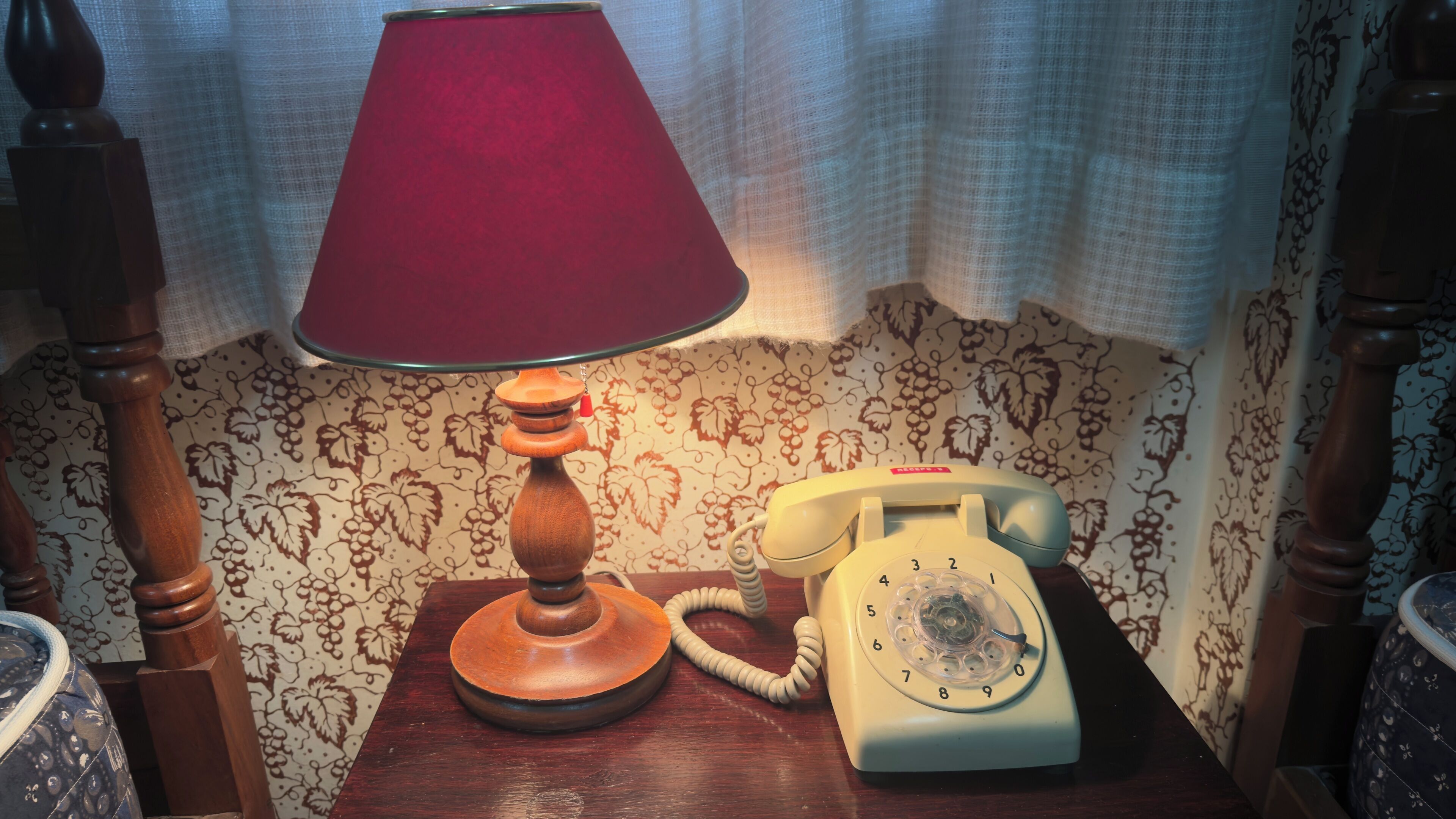 Una mesita de noche de madera con una lámpara roja, un teléfono giratorio blanco y un fondo de papel tapiz estampado.