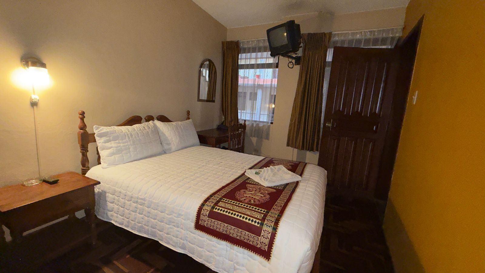 A hotel room with a bed, a bedside table, a television mounted on the wall, and a window with curtains.