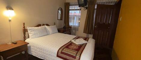 A hotel room with a bed, a bedside table, a television mounted on the wall, and a window with curtains.