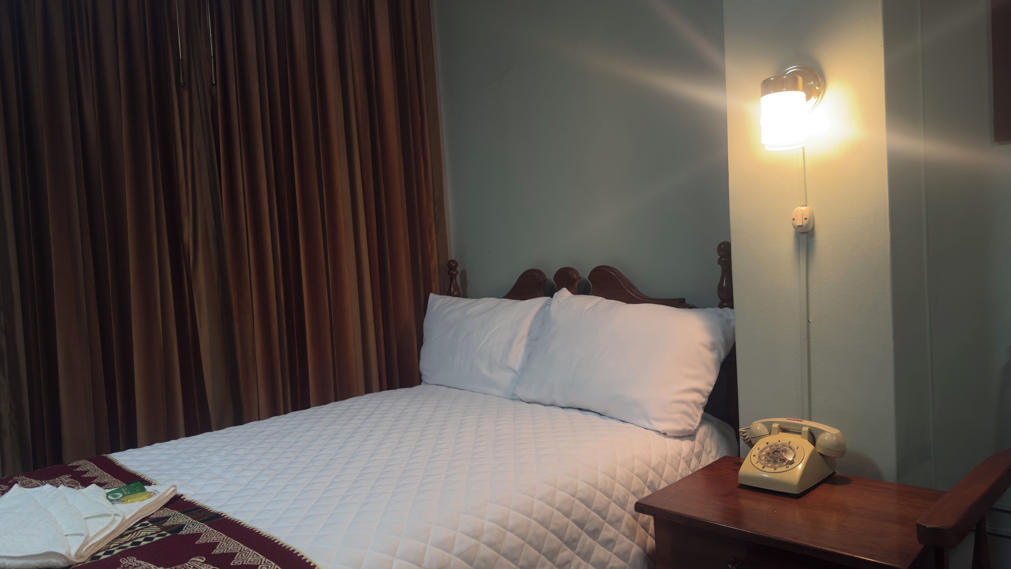 A bed with a quilted white cover, a wooden headboard, a bedside telephone, and a wall-mounted light fixture.