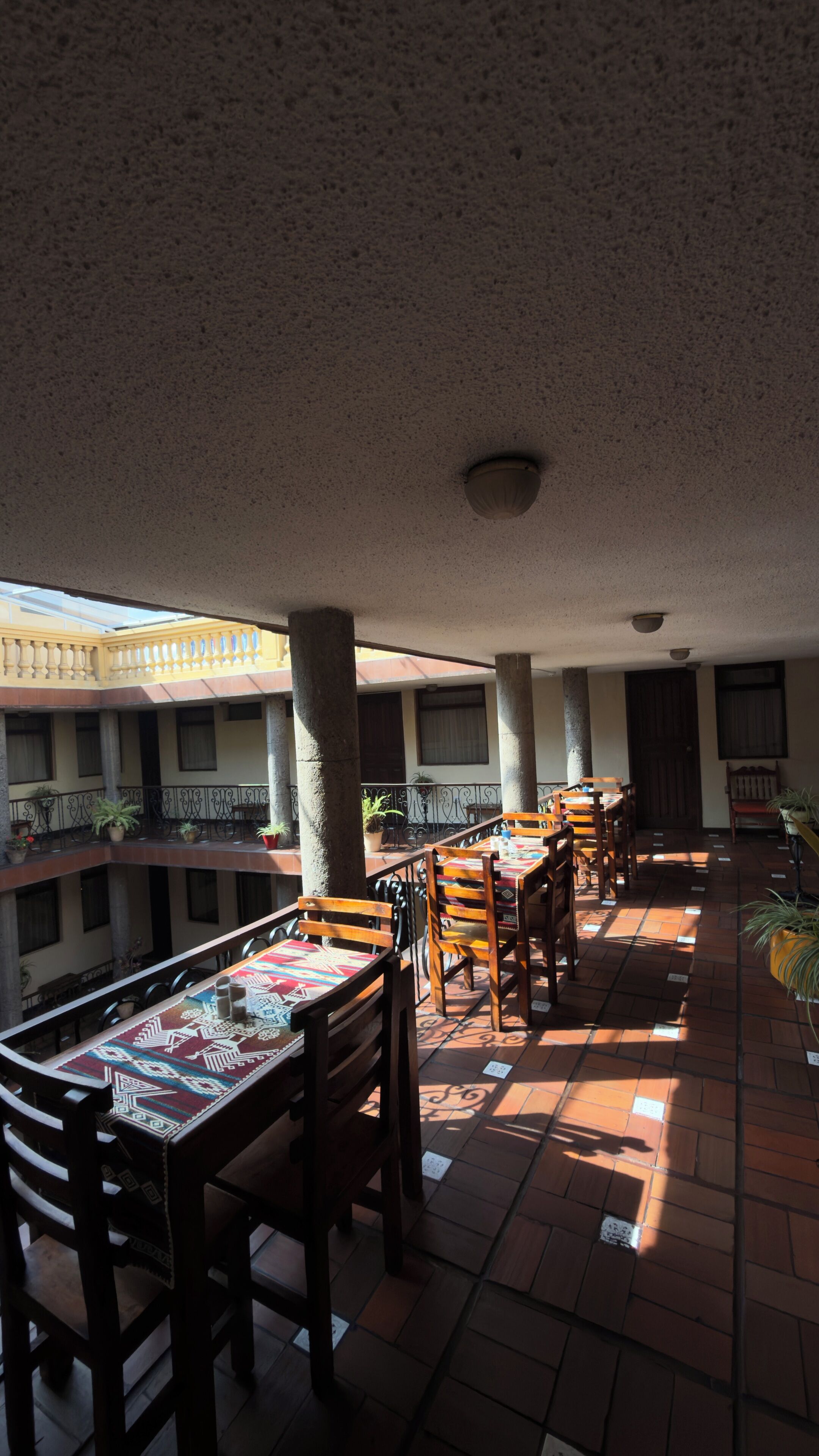 An indoor dining area with wooden tables and chairs, potted plants, and a tiled floor.