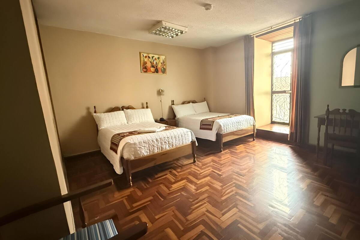 A room with two beds, wooden flooring, a window with curtains, a chair, and a painting on the wall.