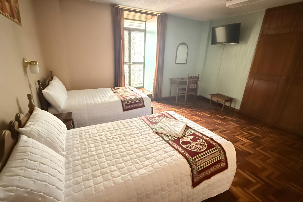 A room with two beds, a wooden floor, a TV mounted on the wall, and a window with curtains.