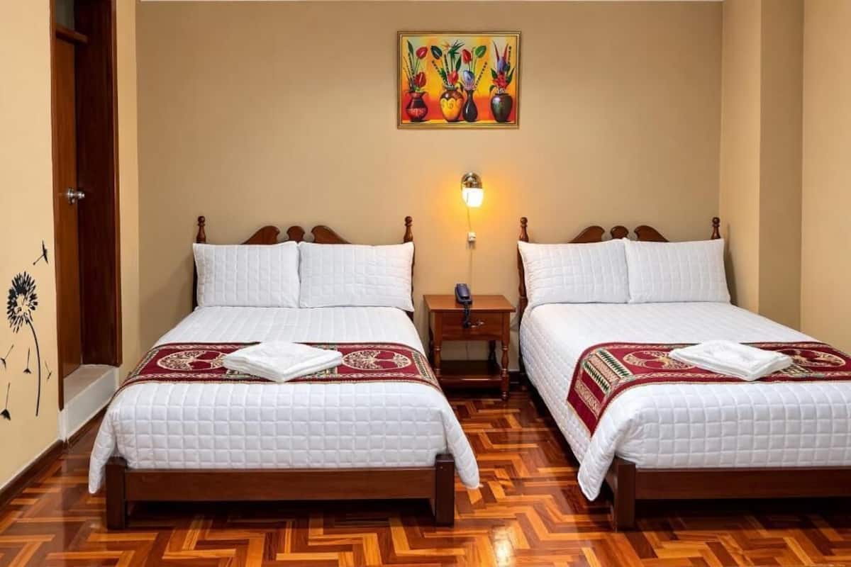 A hotel room with two beds, wooden headboards, a painting above the beds, a bedside table with a lamp, and a wooden floor.