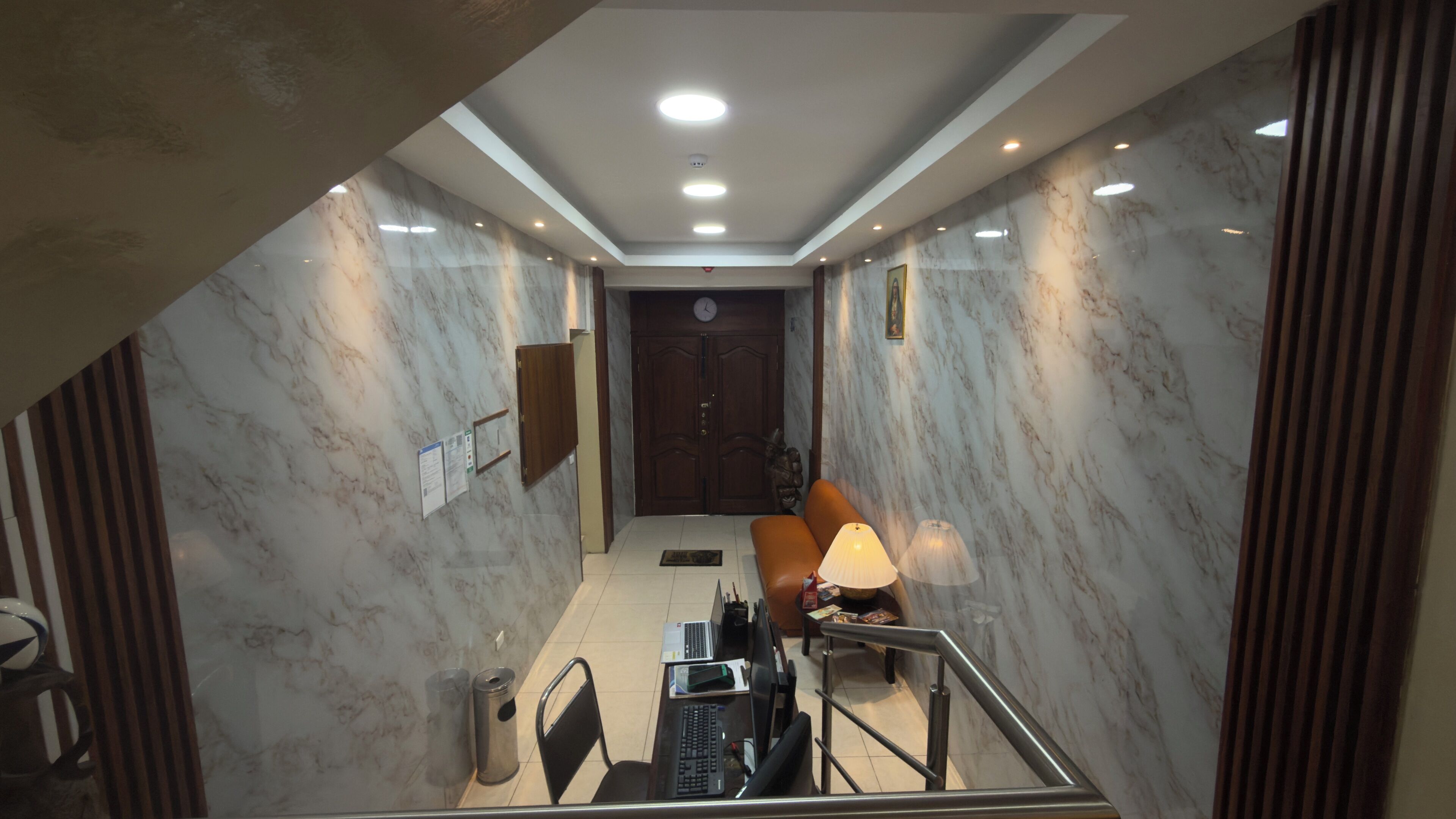 A hotel lobby with marble walls, a wooden door, a seating area with a lamp, and a staircase.