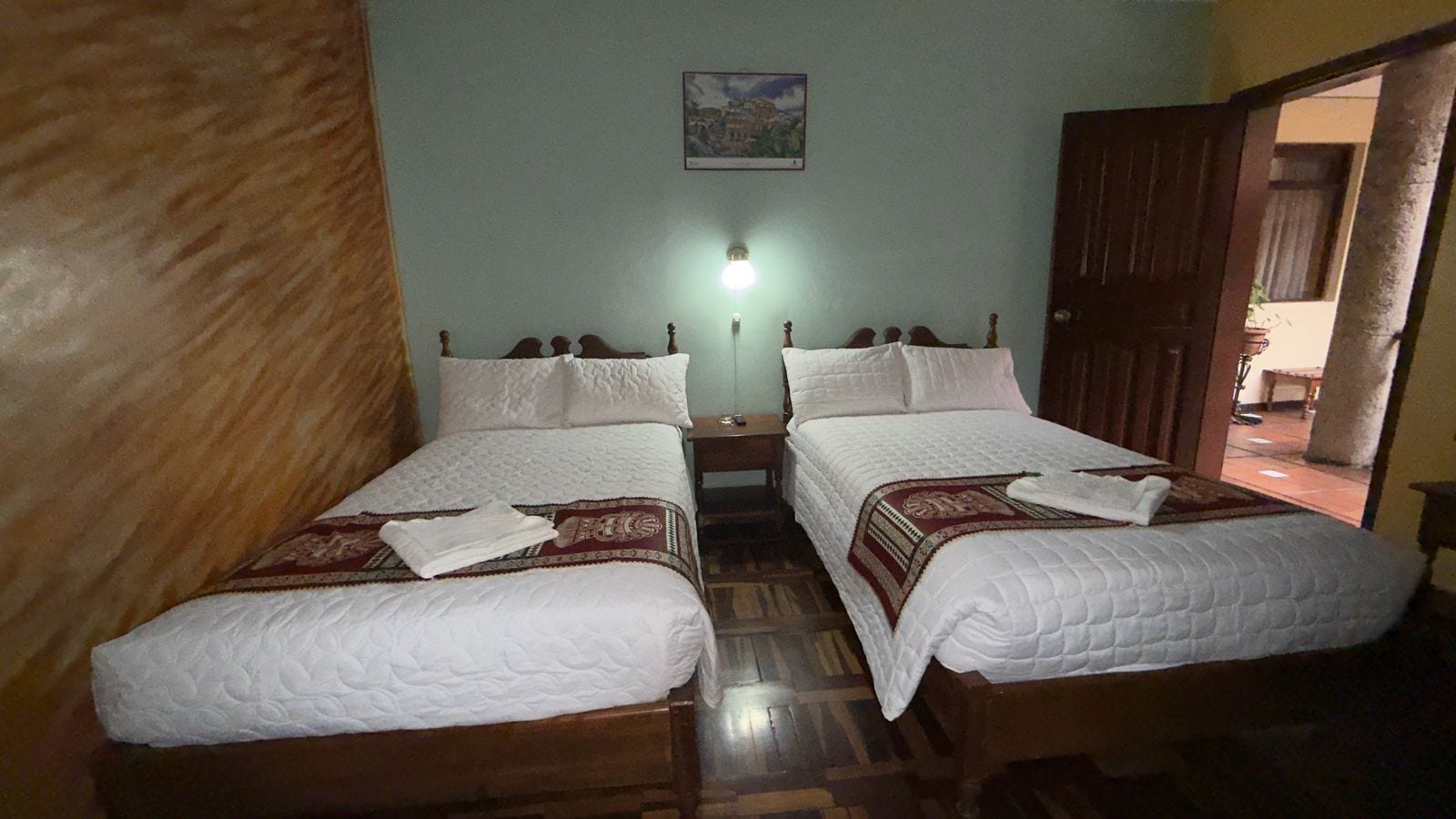 A room with two beds, wooden flooring, a nightstand, a wall-mounted light, and a door leading to another room.
