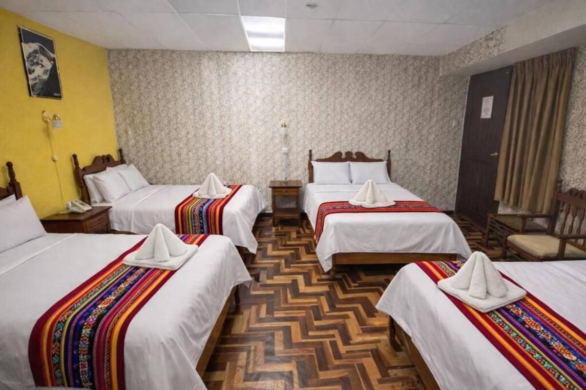 A hotel room with four beds, each with a white bedspread and a colorful blanket. There is a wooden floor, a wooden headboard on each bed, and a wooden chair in the corner.