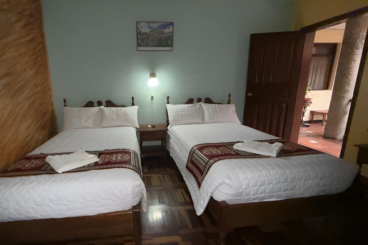 A room with two wooden beds, white bedding, a nightstand with a lamp, and a framed picture on the wall.