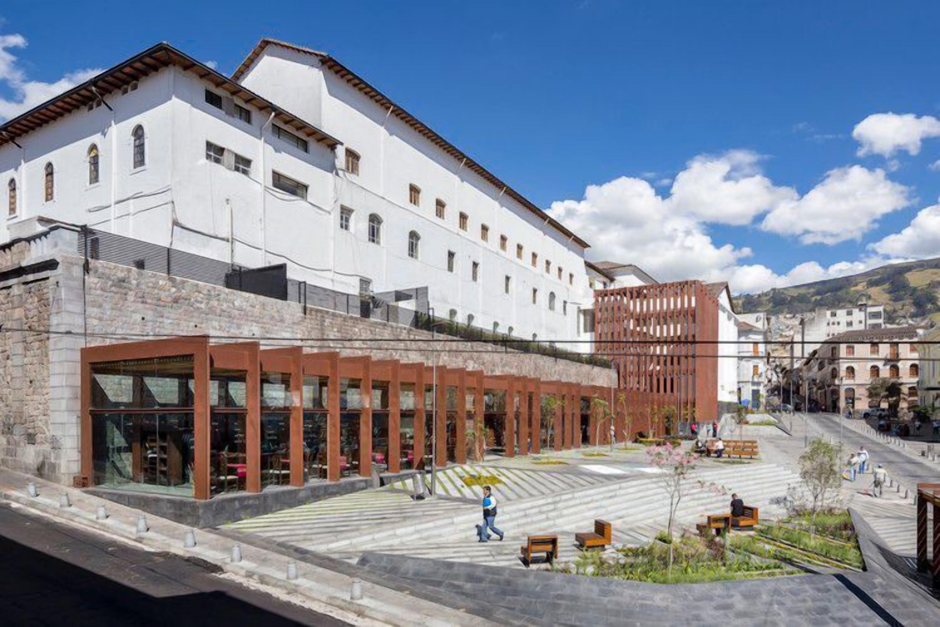 A modern building with a white facade, large windows, and a courtyard with benches and greenery.