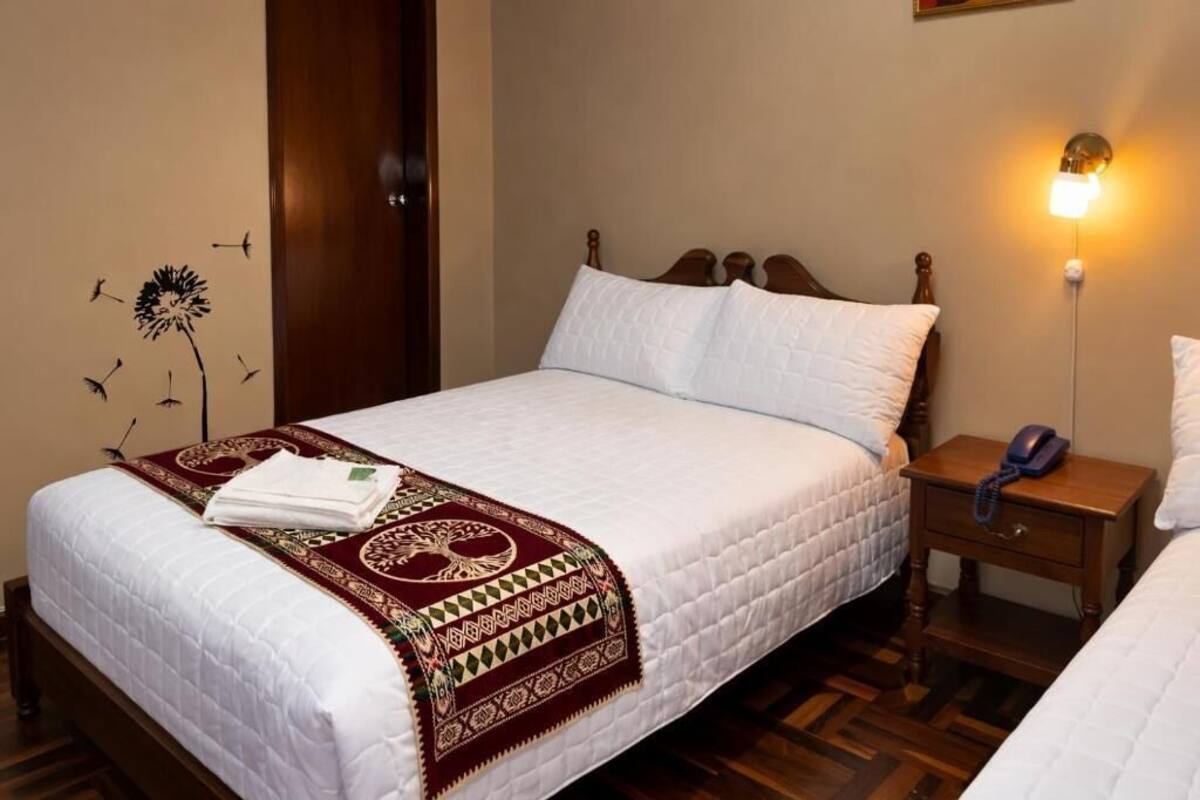 A neatly made bed with a white quilt, a patterned throw blanket, and a wooden nightstand with a lamp.