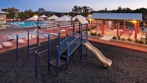 Un parque infantil con tobogán y estructuras de escalada, piscina y un pabellón cubierto con bancos.